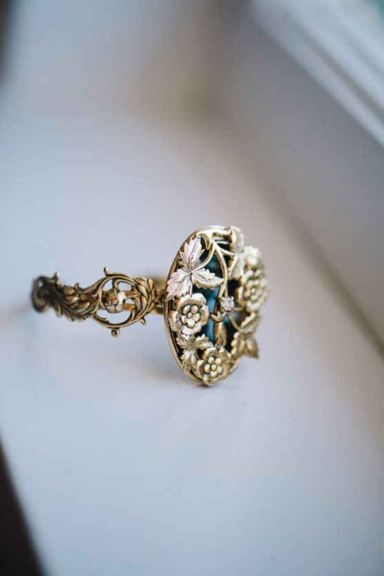 A gold bracelet with intricate floral designs and small pearl accents, featuring an elaborate, round centerpiece with leaves and flowers, placed on a white surface near a window. by Justin Salem Meyer