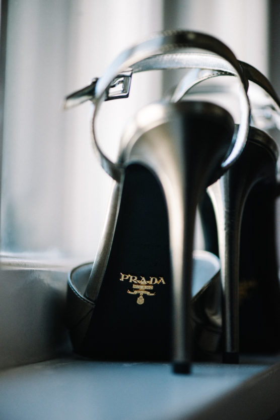 Close-up of a pair of high-heeled silver Prada sandals placed on a windowsill, with the Prada logo visible on the insole of one shoe. by Justin Salem Meyer