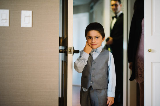 A young boy in a gray vest and bow tie stands in a doorway, smiling and touching his cheek, with two adults blurred in the background. by Justin Salem Meyer