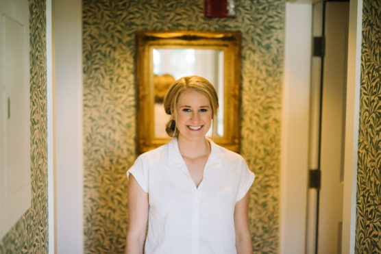 A woman with blonde hair, wearing a white shirt, stands smiling in a hallway with patterned green wallpaper and a gold-framed mirror behind her. by Justin Salem Meyer