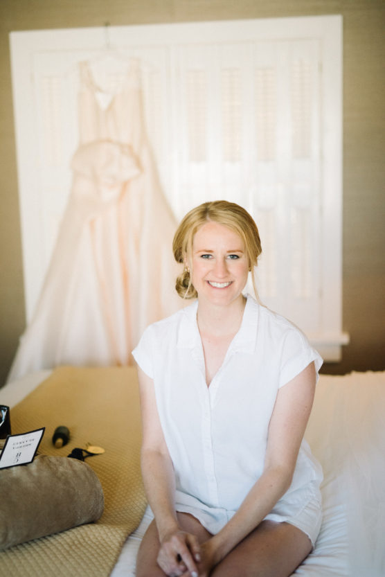 A woman with blond hair in a loose updo, wearing a white button-up shirt, sits smiling on a bed. Behind her, a white dress hangs on a door. The room is softly lit and cozy. by Justin Salem Meyer