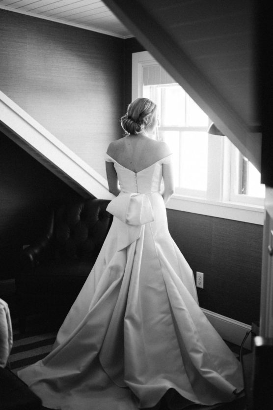 A woman in an elegant, off-the-shoulder wedding dress stands indoors by a window, facing outside. The photo is in black and white, highlighting the gown’s full skirt and large bow at the back. by Justin Salem Meyer