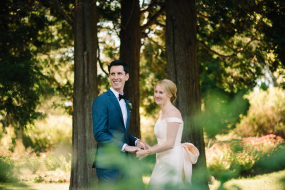 A bride in an off-shoulder white dress and a groom in a blue suit smile and hold hands outdoors, standing in front of tall trees with greenery and sunlight in the background. by Justin Salem Meyer