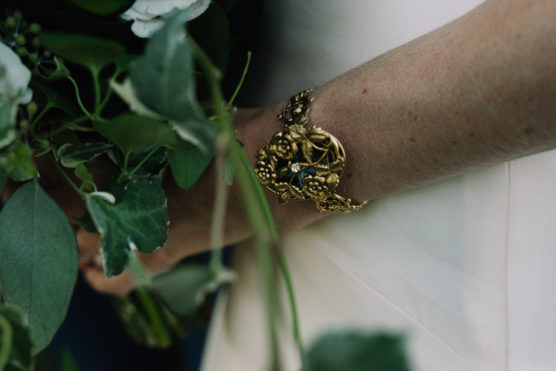 A close-up of a persons arm wearing an ornate gold bracelet with floral designs, holding a bouquet of green leaves and white flowers. The person is dressed in a light-colored garment. by Justin Salem Meyer