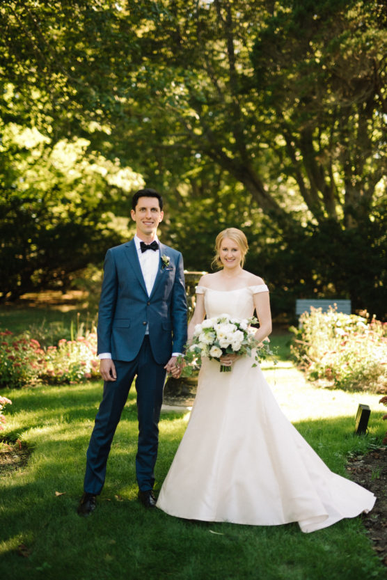 A bride in an off-the-shoulder white gown holds a bouquet of flowers while standing next to a groom in a blue suit and bow tie. They smile in a sunlit garden surrounded by greenery. by Justin Salem Meyer