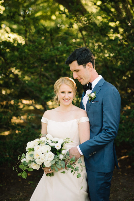 A bride in an off-shoulder white dress holds a bouquet of white flowers and greenery, standing next to a groom in a blue suit and bow tie, with greenery and sunlight in the background. by Justin Salem Meyer