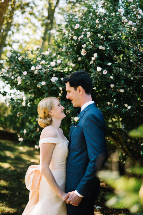 A bride and groom stand outdoors, holding hands and smiling at each other in front of a lush, flowering bush. The bride wears an off-shoulder white dress; the groom is in a blue suit. Sunlight filters through the trees behind them. by Justin Salem Meyer