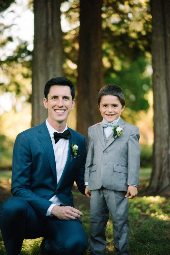 A man in a blue suit and bow tie kneels next to a young boy in a light gray suit. Both are smiling and wearing boutonnieres, and they are outdoors with tall trees in the background. by Justin Salem Meyer