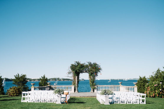 An outdoor wedding setup features rows of white chairs facing a floral archway on a lawn overlooking a blue body of water, with sailboats and a clear sky in the background. by Justin Salem Meyer