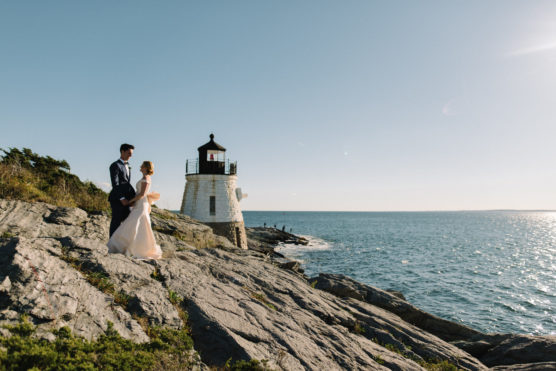 A bride and groom stand on rocky cliffs beside a lighthouse, overlooking the ocean on a clear, sunny day. The bride wears a flowing gown, and the groom is in a dark suit, both facing the water. by Justin Salem Meyer