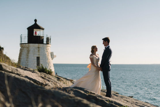 A bride and groom stand on rocky ground near a lighthouse, holding hands and gazing at each other, with the ocean and clear sky in the background. by Justin Salem Meyer