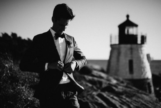 A man in a suit adjusts his jacket while standing outdoors near a lighthouse. The black and white image creates a dramatic, classic atmosphere. by Justin Salem Meyer