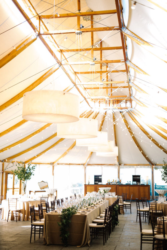 A bright, elegant tented event space with long banquet tables set for a meal, wooden chairs, greenery centerpieces, large hanging lights, and natural sunlight streaming in. by Justin Salem Meyer