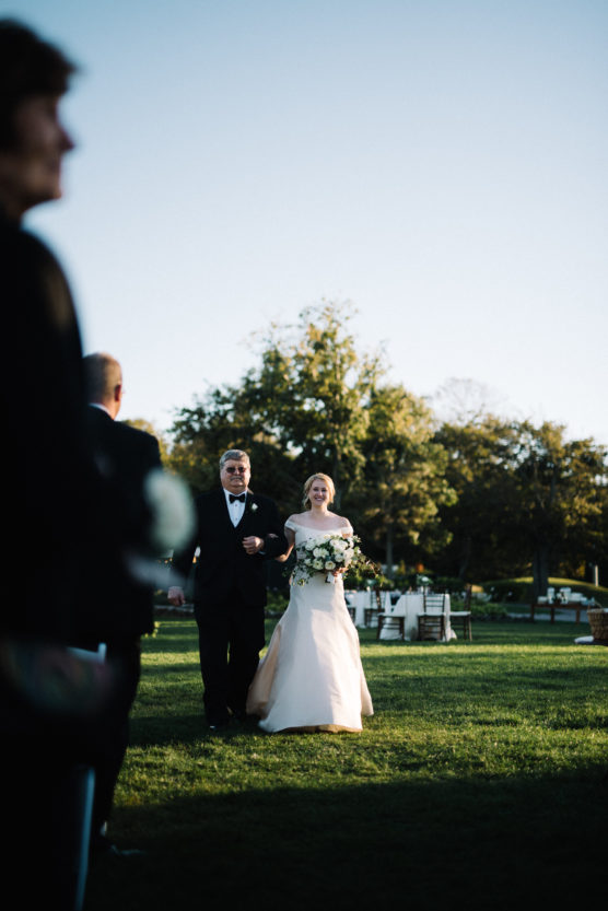 A bride in a white gown holding a bouquet walks arm-in-arm with a man in a tuxedo across a grassy outdoor area, with guests and trees in the background under a clear sky. by Justin Salem Meyer