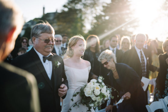 A bride in a white dress smiles while walking down the aisle with an older man in a tuxedo at an outdoor wedding, surrounded by guests in formal attire and sunlight streaming in. by Justin Salem Meyer