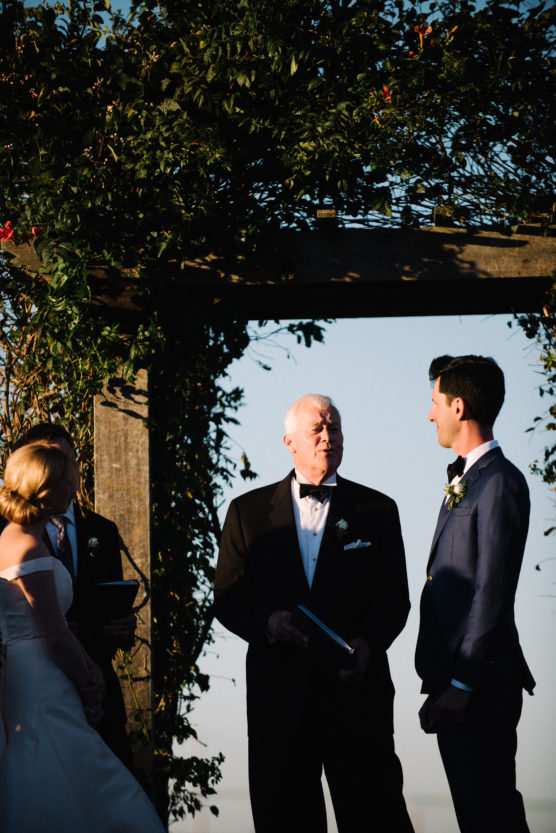 A bride and groom stand with an officiant under a wooden arch covered in greenery during an outdoor wedding ceremony, with sunlight casting shadows on them. by Justin Salem Meyer
