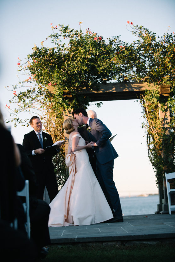 A bride and groom share a kiss under a flower-covered arch outdoors by the water as an officiant smiles nearby, during their wedding ceremony. by Justin Salem Meyer