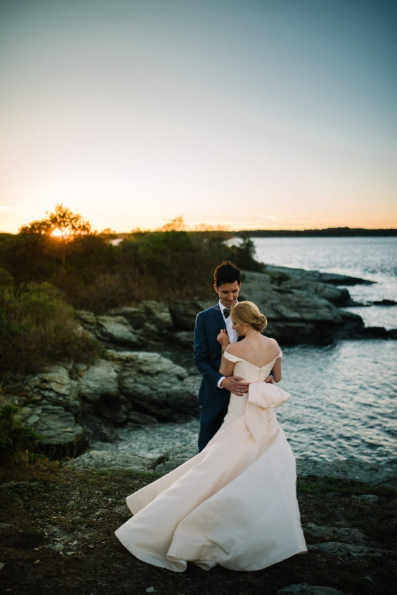 A bride and groom embrace near rocky shores at sunset, with the bride in a white dress and the groom in a blue suit. The sun sets over the water in the background, creating a romantic atmosphere. by Justin Salem Meyer