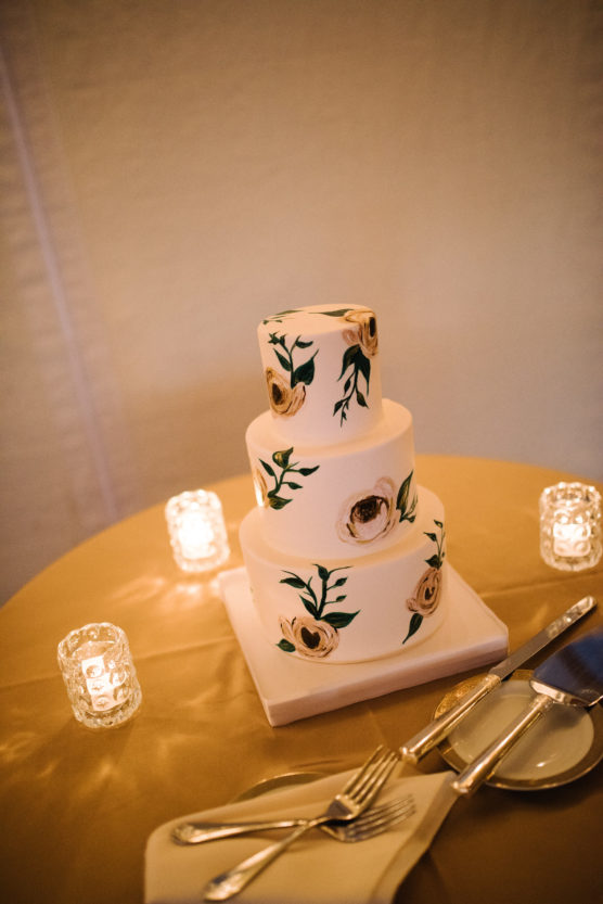 A three-tier white cake with hand-painted floral designs sits on a round, golden tablecloth, surrounded by three small lit candles, with utensils and a napkin neatly arranged nearby. by Justin Salem Meyer