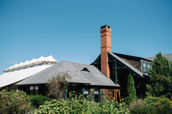 A rustic house with a brick chimney and shingled roof sits among green bushes and trees under a clear blue sky, with a large white event tent visible in the background. by Justin Salem Meyer