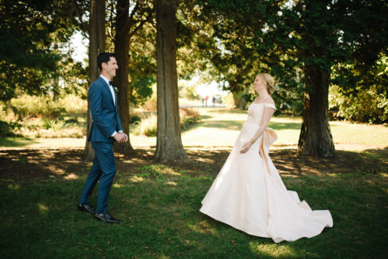 A bride in a flowing white dress and a groom in a blue suit smile joyfully at each other outdoors, surrounded by tall trees and greenery, on a sunny day. by Justin Salem Meyer