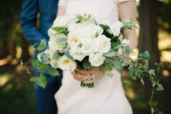 A bride in a white dress holds a large bouquet of white roses and greenery. A person in a blue suit stands beside her, partially visible. The background is blurred greenery outdoors. by Justin Salem Meyer