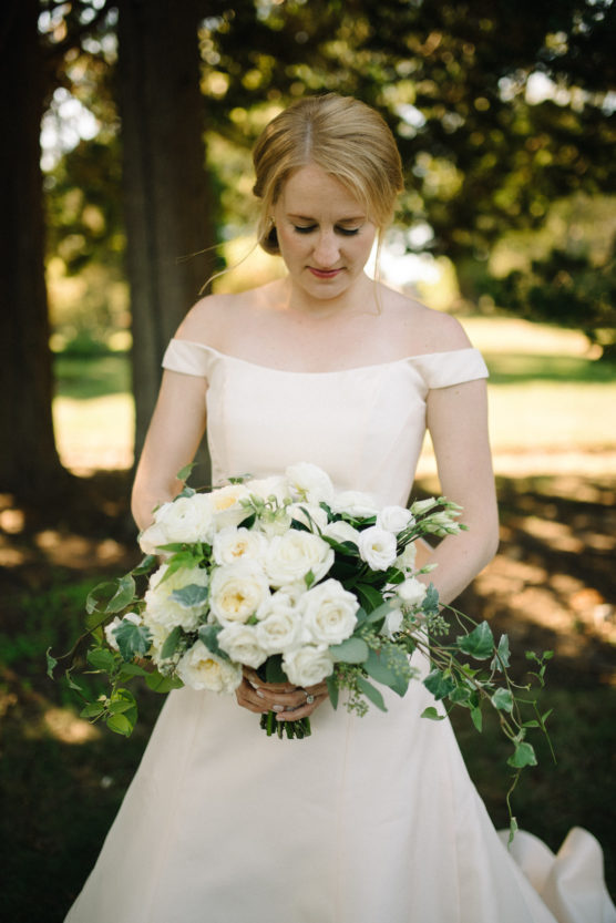 A bride in an off-shoulder white dress stands outdoors, holding a large bouquet of white roses and greenery, with sunlight filtering through trees in the background. by Justin Salem Meyer