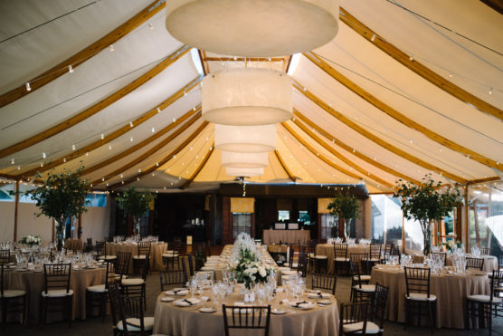 A decorated event tent features round tables with tan tablecloths, elegant place settings, floral centerpieces, wooden chairs, large hanging lanterns, and string lights, creating a festive and formal atmosphere. by Justin Salem Meyer