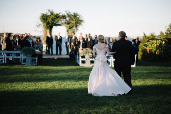 A bride in a white dress walks across a grassy lawn with an older man towards a group of seated guests and a floral arch at an outdoor wedding ceremony. by Justin Salem Meyer