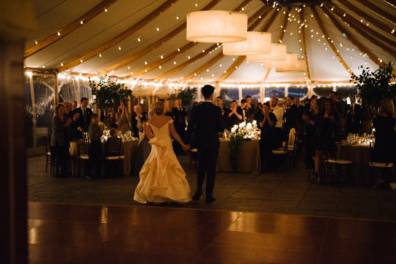 A bride and groom, holding hands, enter a warmly lit wedding reception tent filled with round tables, guests standing and applauding, and string lights hanging from the ceiling. by Justin Salem Meyer