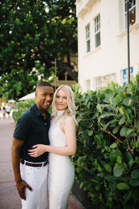 A smiling couple poses together on a sidewalk next to green bushes and a light-colored building. The man wears a dark shirt and white pants; the woman wears a white dress. Trees and people are visible in the background. by Justin Salem Meyer