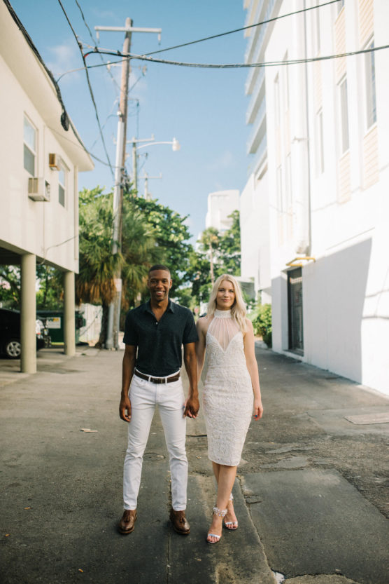 A couple holds hands and smiles while standing in a sunny urban alley. The man wears a dark shirt and white pants, and the woman wears a white dress and heels. Tall buildings and power lines are in the background. by Justin Salem Meyer