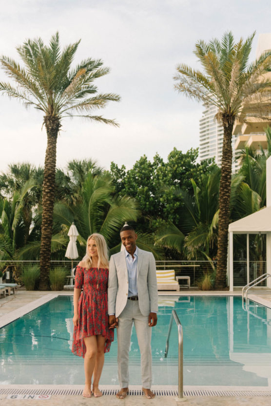 A couple stands hand in hand at the edge of a pool, surrounded by palm trees. The woman wears a red floral dress, and the man is dressed in a light grey suit. Modern buildings are visible in the background. by Justin Salem Meyer