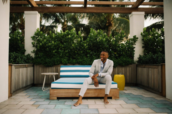 A man in a light gray suit sits barefoot on a striped outdoor lounge chair, smiling. He is surrounded by lush greenery and palm trees on a tiled patio with wooden pergola overhead. by Justin Salem Meyer