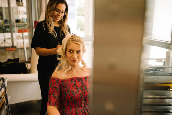 A woman in a red off-the-shoulder dress sits smiling while a hairstylist styles her blonde hair in a bright, modern salon. by Justin Salem Meyer
