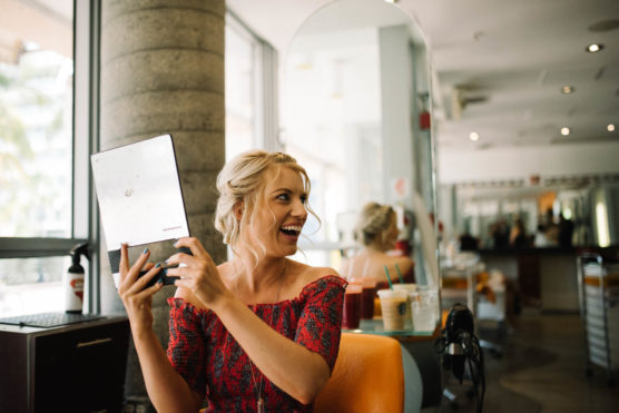 A smiling woman with blonde hair sits in a salon chair, holding up a mirror and looking at her reflection. She wears a red patterned off-shoulder top, and drinks are on the table beside her. The salon is bright and modern. by Justin Salem Meyer