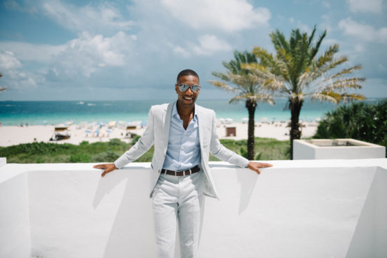 A man in a light gray suit and sunglasses smiles while standing on a white terrace overlooking a tropical beach with palm trees, blue ocean, and cloudy sky in the background. by Justin Salem Meyer