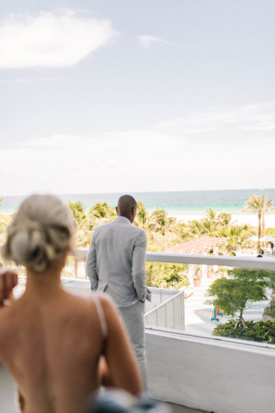 A man in a light gray suit stands on a balcony overlooking a tropical beach, with palm trees and ocean in the background. A woman with blonde hair in an updo stands in the foreground, slightly out of focus. by Justin Salem Meyer
