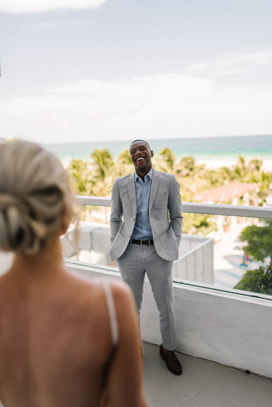 A man in a light gray suit stands on a balcony, smiling with hands in pockets, facing a woman with blonde hair in an updo. The background shows palm trees, the ocean, and a partly cloudy sky. by Justin Salem Meyer