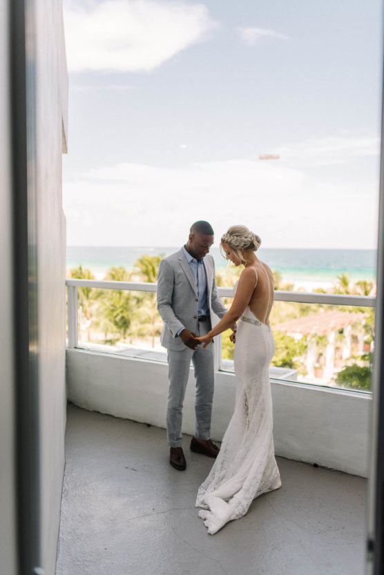 A bride and groom stand on a balcony overlooking palm trees and the ocean, holding hands. The bride wears a backless white gown and the groom is in a light gray suit. They face each other in an intimate moment. by Justin Salem Meyer