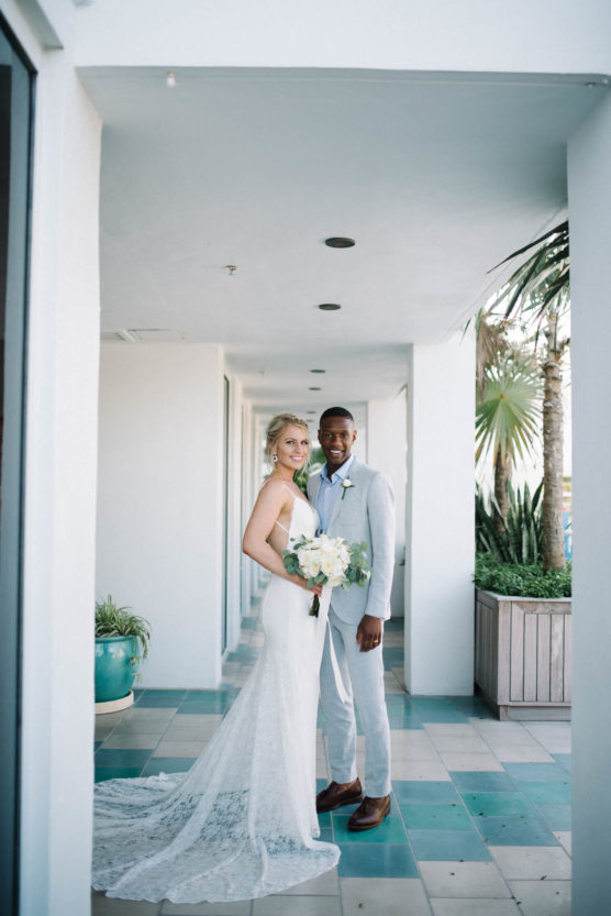 A bride in a white dress and a groom in a light gray suit stand together in a sunny, modern hallway with potted plants, smiling and holding a bouquet of white flowers. by Justin Salem Meyer