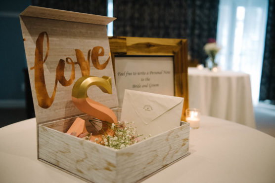 A wooden box with love on the lid holds a large decorative letter “S,” an envelope, and white flowers. A framed sign and a lit candle sit beside the box on a round table at an event. by Justin Salem Meyer