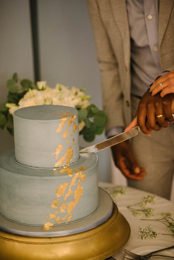 A couple cuts a two-tier blue cake with gold accents together; their hands, one with a ring, hold a knife. The scene is decorated with white flowers and greenery. by Justin Salem Meyer