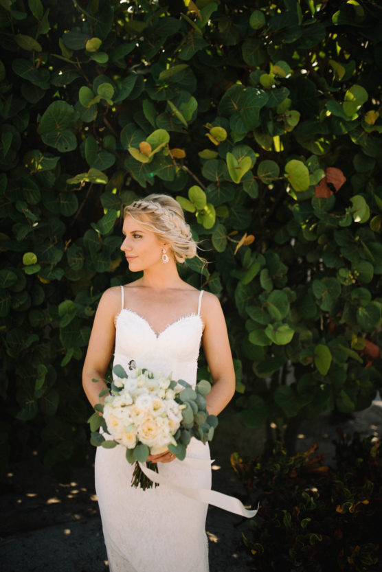 A bride in a white lace dress holding a bouquet of white flowers stands in front of lush green foliage, looking to the side with her hair in a braided updo. by Justin Salem Meyer