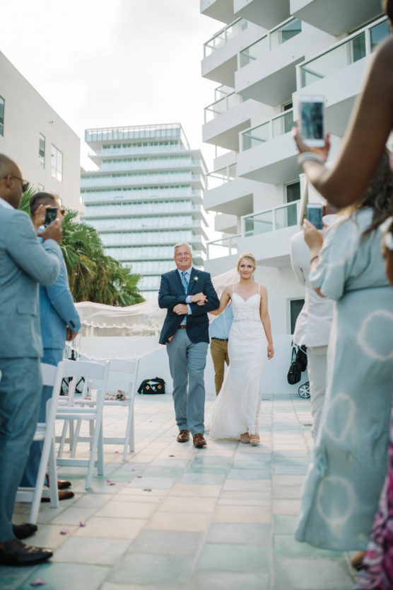 A bride in a white dress walks down an outdoor aisle with an older man, smiling, as guests in formal attire stand and take photos at a modern venue with white buildings. by Justin Salem Meyer