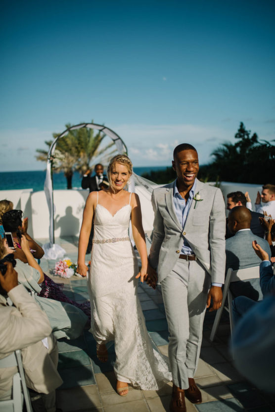 A newlywed couple, smiling and holding hands, walk down the aisle outdoors at a sunny seaside wedding with guests applauding on either side. by Justin Salem Meyer