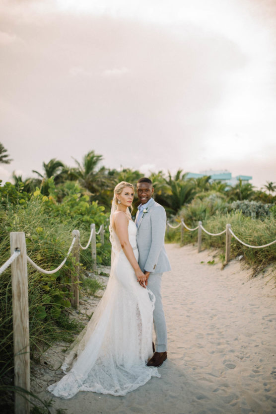 A bride in a white lace gown and a groom in a light gray suit stand hand in hand on a sandy path lined with greenery, smiling at the camera with palm trees and a pastel sky in the background. by Justin Salem Meyer