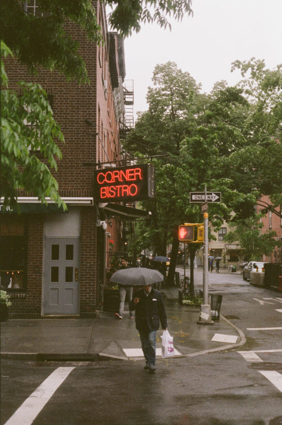 A person with an umbrella crosses a wet street corner near a brick building with a red neon Corner Bistro sign. Trees and a traffic light are visible on a cloudy day. by Justin Salem Meyer