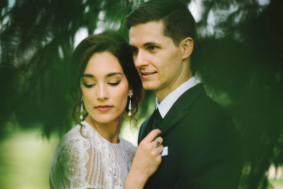 A bride and groom pose closely together outdoors, surrounded by greenery. The bride, wearing lace and earrings, gently holds the grooms lapel and closes her eyes, while the groom in a suit looks ahead and smiles softly. by Justin Salem Meyer