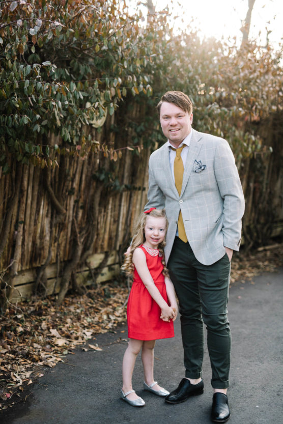A man in a light plaid blazer and yellow tie stands smiling next to a young girl in a red dress and silver shoes. They are outdoors on a path with fallen leaves and a wooden fence behind them. by Justin Salem Meyer
